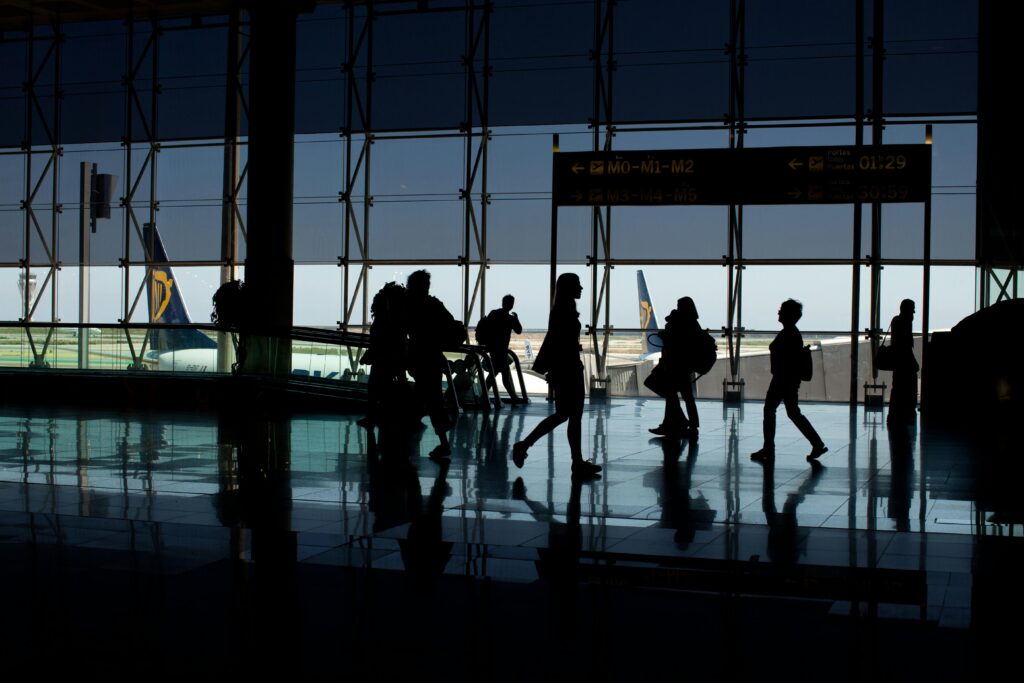 Silhouettes of people walking in an airport terminal, plane tail visible outside.